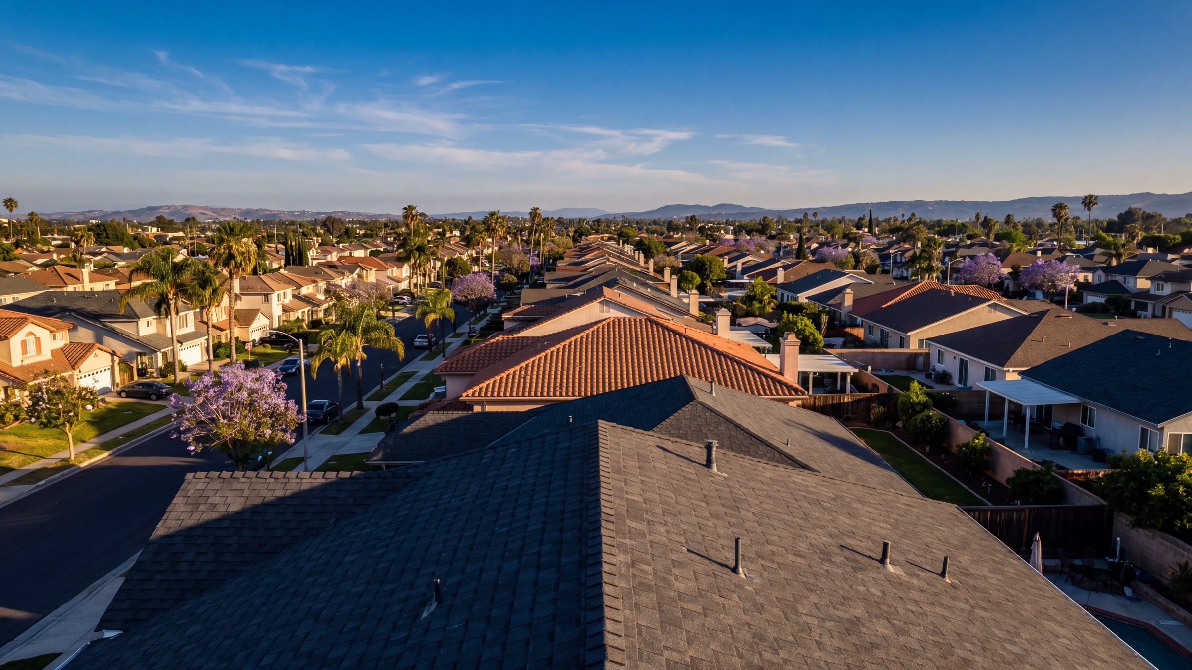 Aerial view of a Southern California neighborhood at golden hour, rows of asphalt shingle and terracotta tile roofs stretching to the horizon